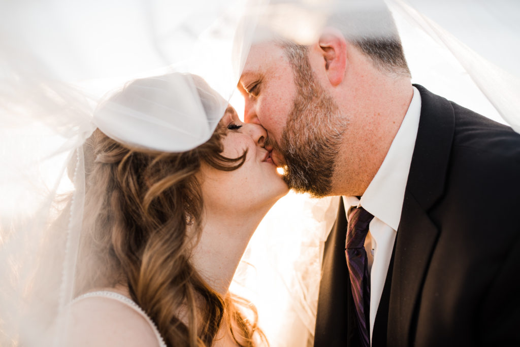 bride and groom kissing under veil with sunlight behind