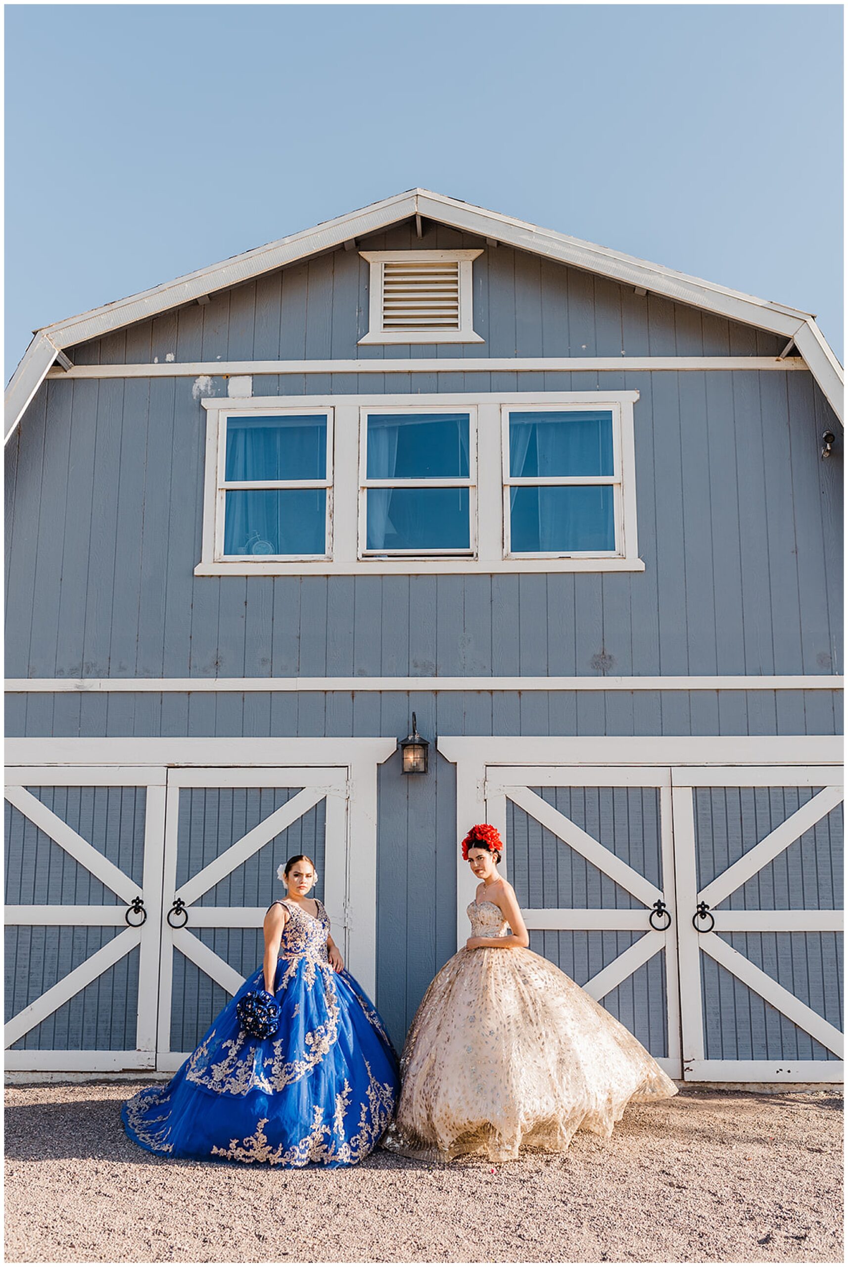 Two teenagers in ornate ball gowns stand in front of a large blue barn Phoenix Quinceaneras dresses