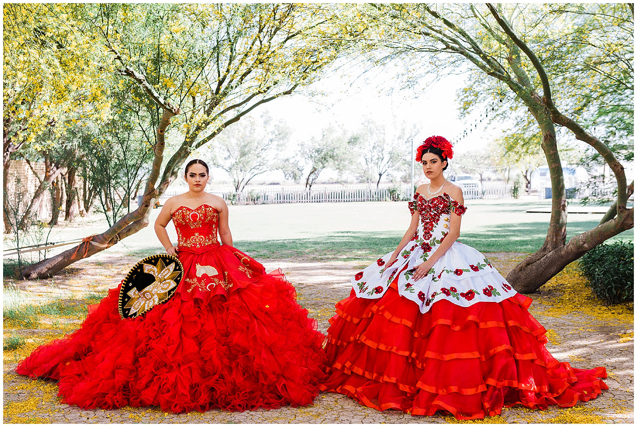 Two teenagers in large Quinceaneras gowns stand under trees with yellow flowers