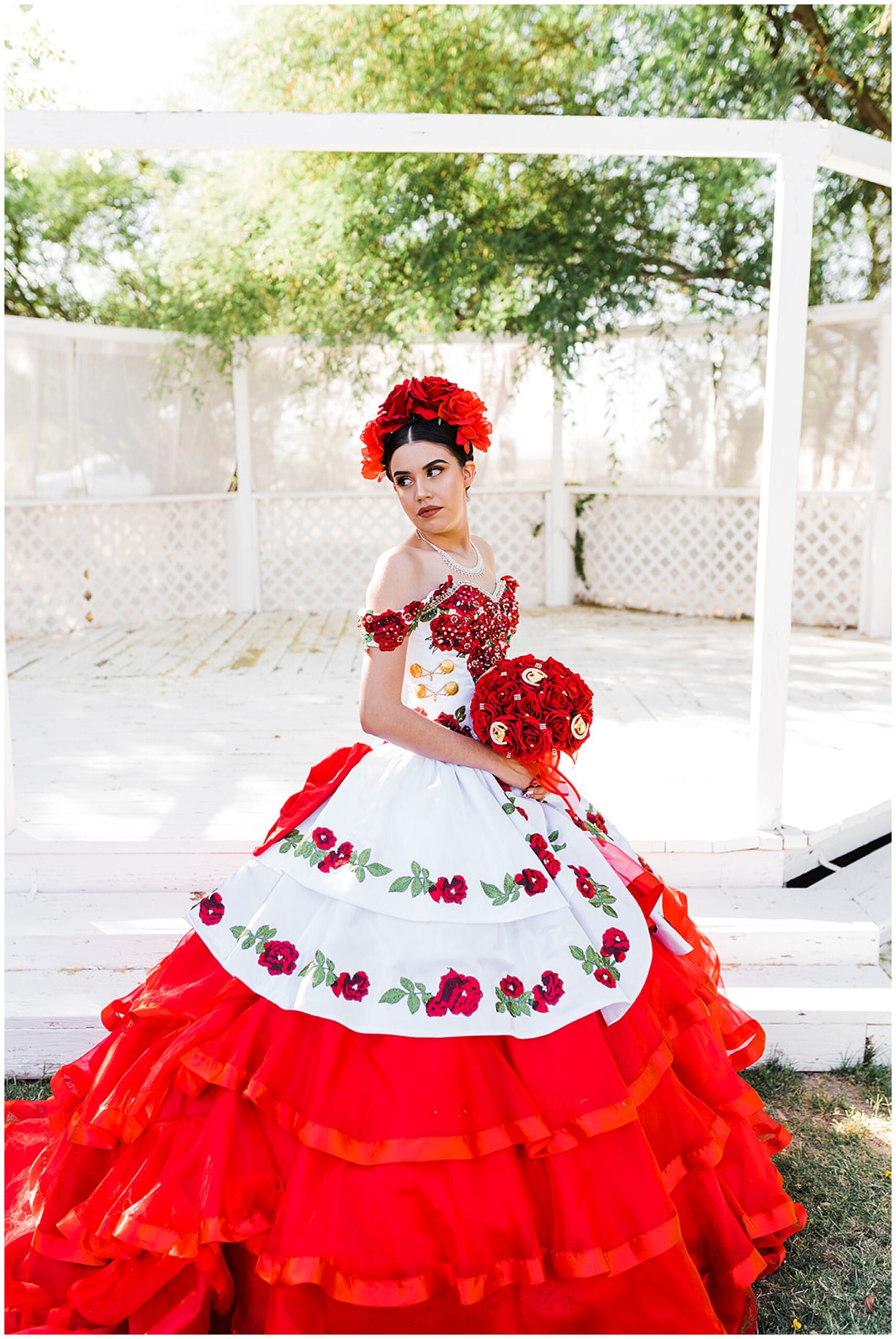 Teenager stands in a red and white ball gown embroidered with red roses while holding a red rose bouquet
