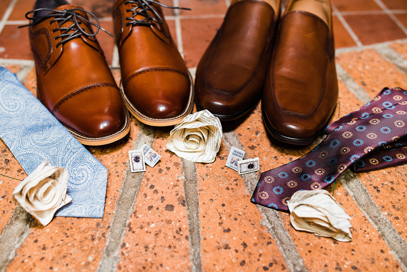 Wedding details of shoes, ties cufflinks and carnations make of pages from a book sit on a brick floor