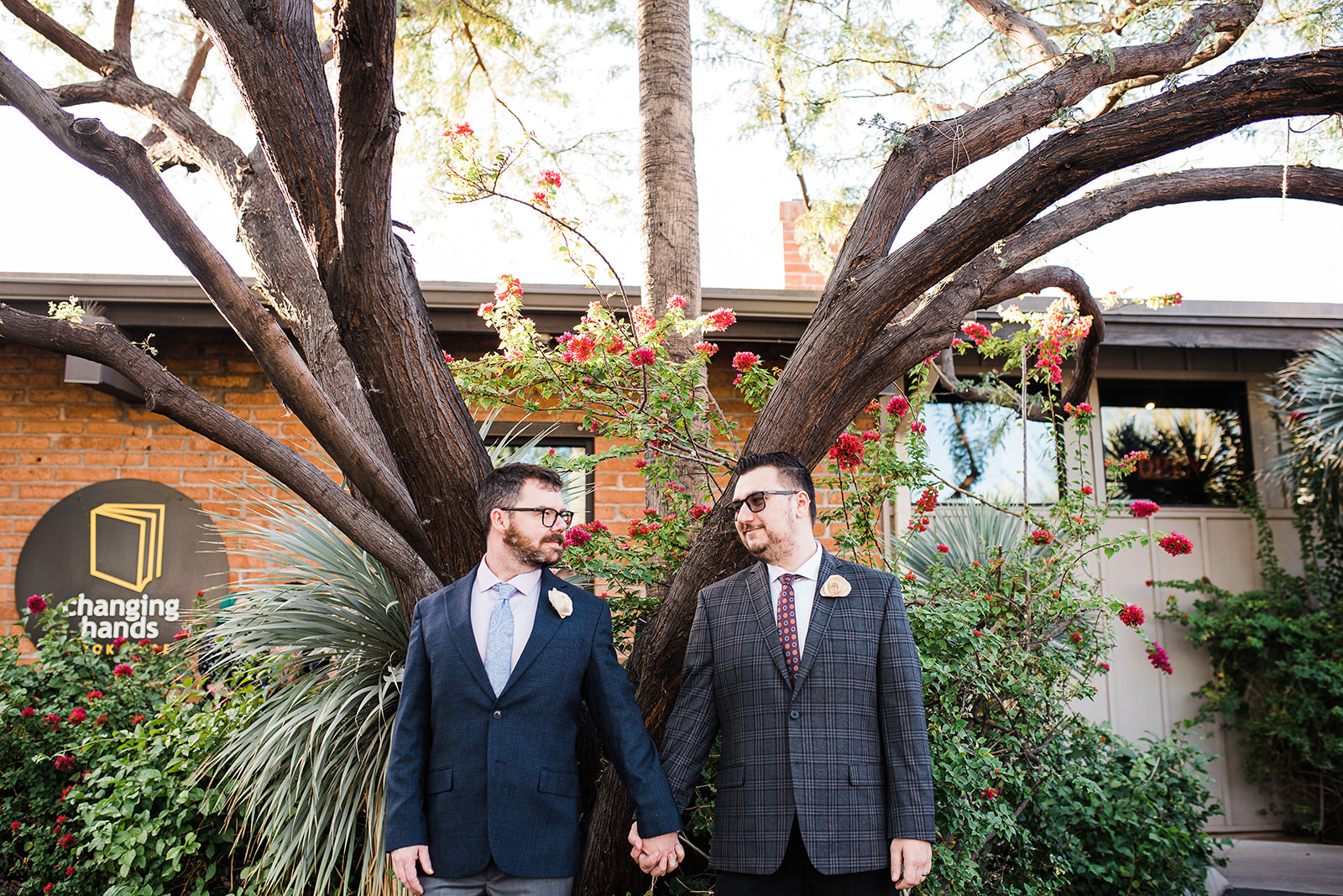 Newlywed men stand in front of a bookstore holding hands under a tree and flowering bush