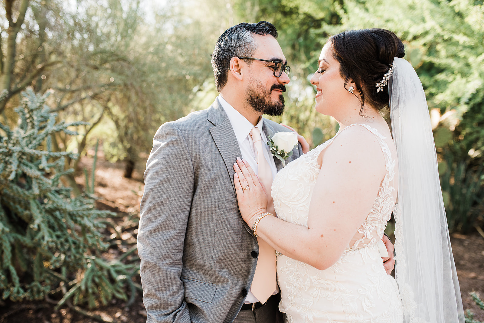 Newlyweds stand nose to nose in a desert garden while the bride wears a white lace dress and long veil at Bella Rose Estate Wedding