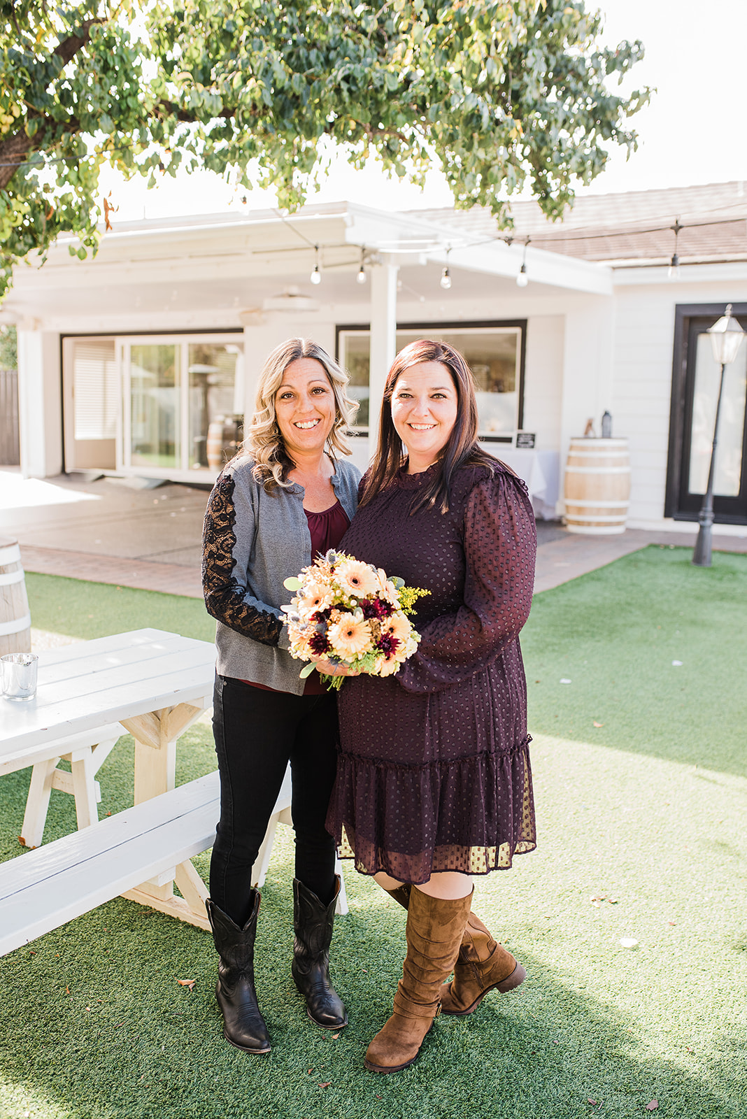 A bride in a purple dress and brown boots stands in the grassy yard under a tree with her maid of honor and a large flowery bouquet at The Cottage Wedding Venue