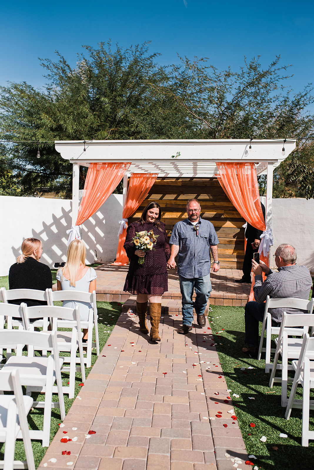 Newlyweds walk down the aisle after their ceremony under a pergola with orange drapes The Cottage Wedding Venue