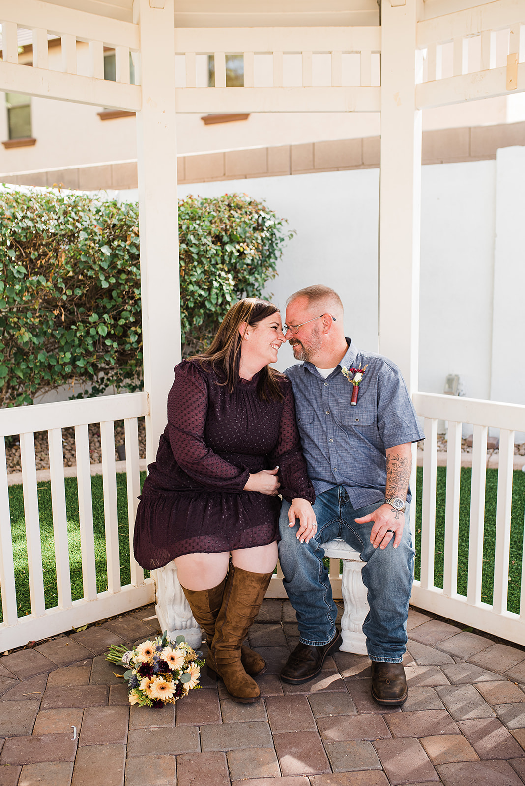 Newlyweds sit on a bench under a gazebo on a stone patio in denim and a purple dress