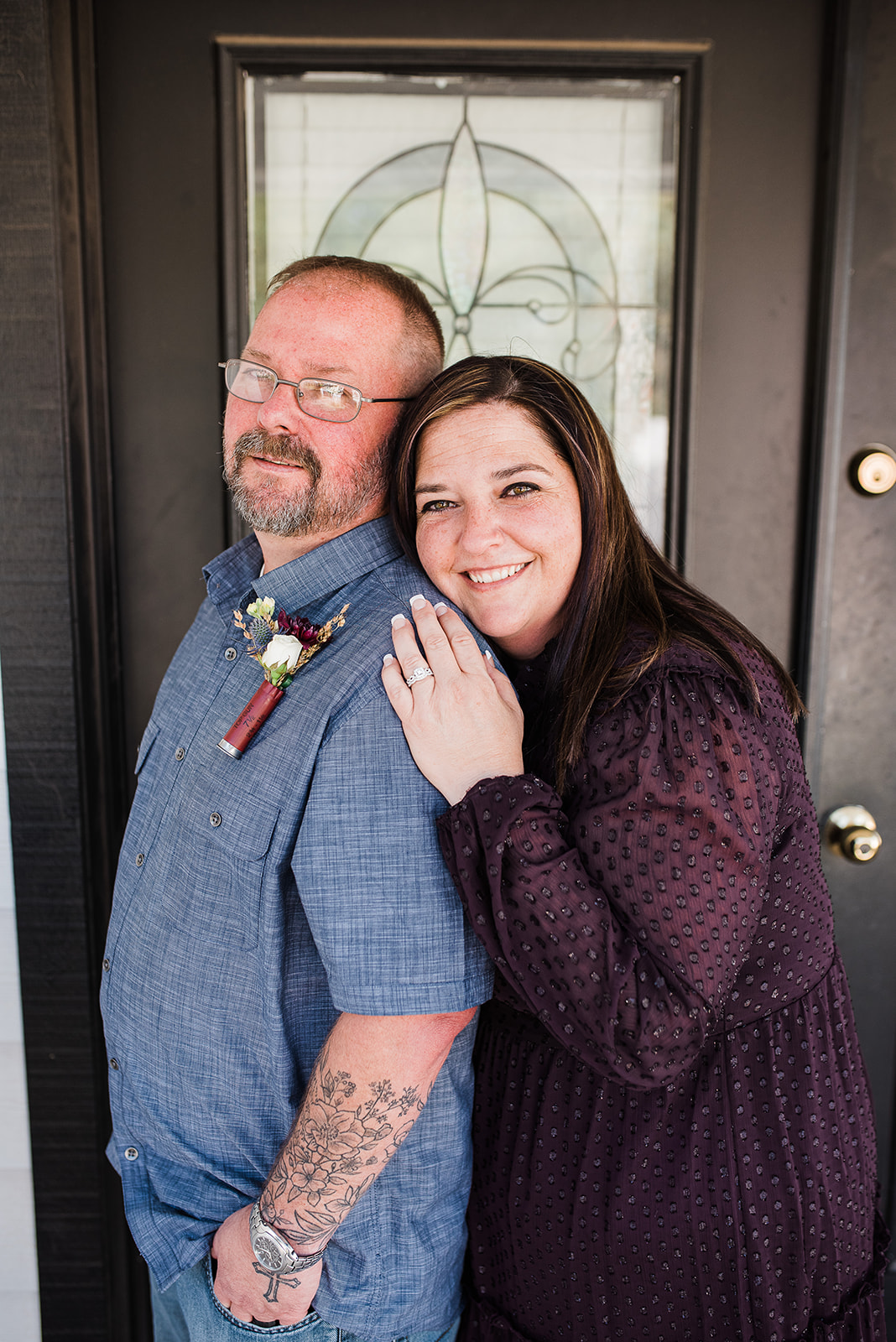 A bride in a purple dress leans on the shoulder of her new husband wearing a denim shirt and purple boutonnière at the front door of The Cottage Wedding Venue
