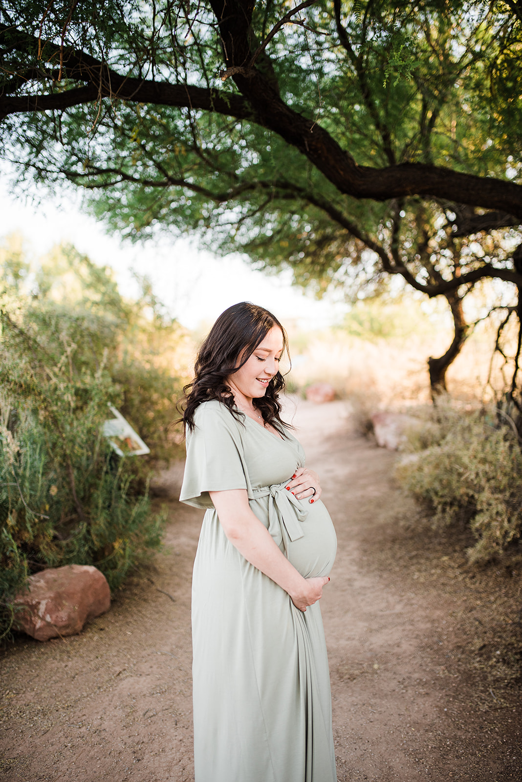 A mother to be smiles down to her bump in her hands while standing in a desert trail in a green maternity dress