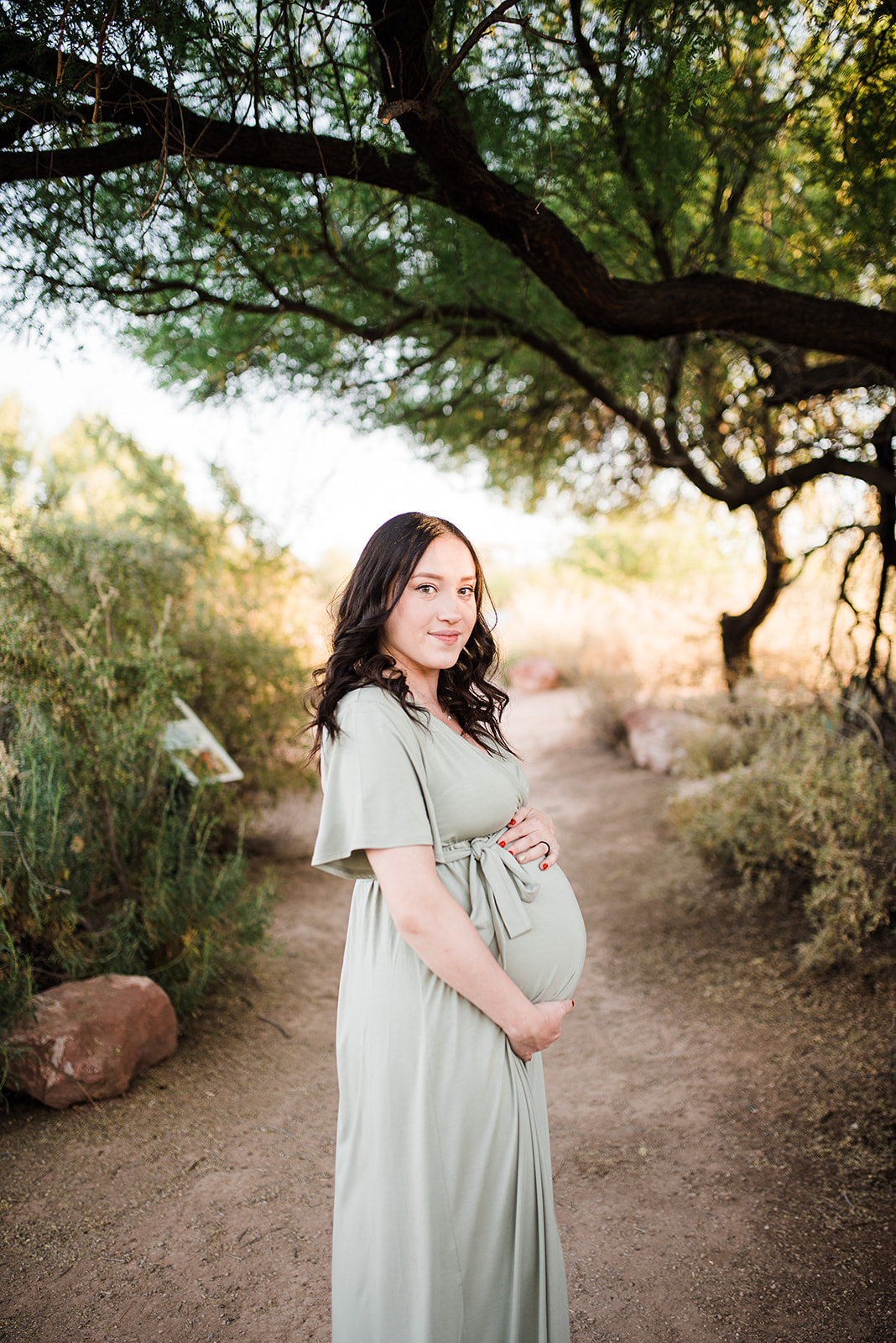 A bride in a sage maternity gown stands in a park trail under a tree at sunset while holding her bump with both hands Phoenix Birth Center