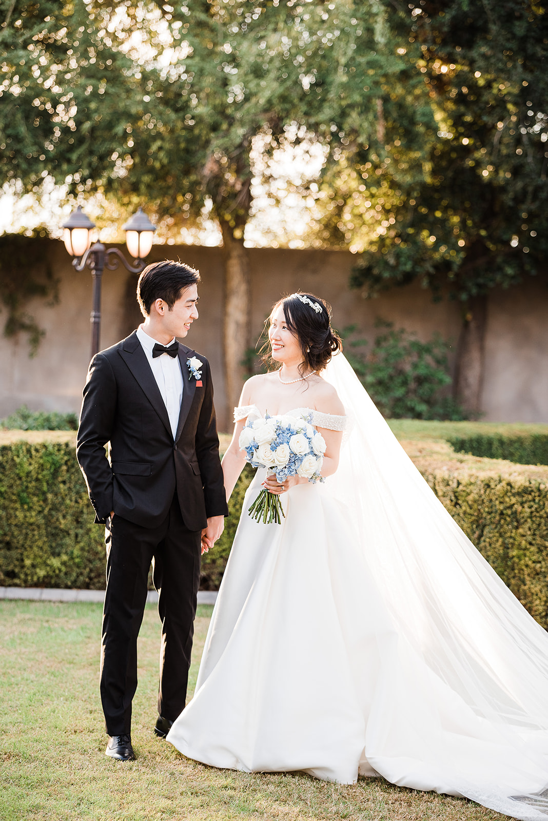 Newlyweds smile at each other in a black tuxedo and white silk dress with long veil and train holding hands in a Phoenix Country Club Weddings venue garden