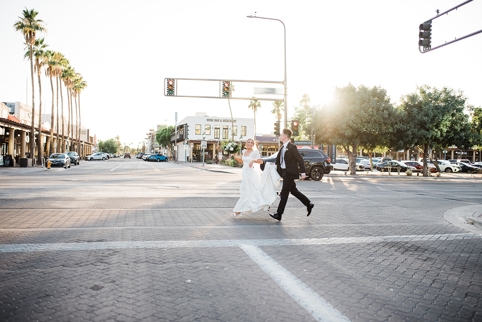 Newlyweds walk across an intersection while the groom holds the train and bride her white flowers