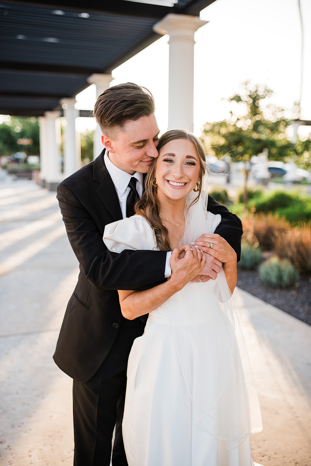 Newlyweds snuggle under a covered walkway in a black suit and white silk dress Phoenix Wedding Planners