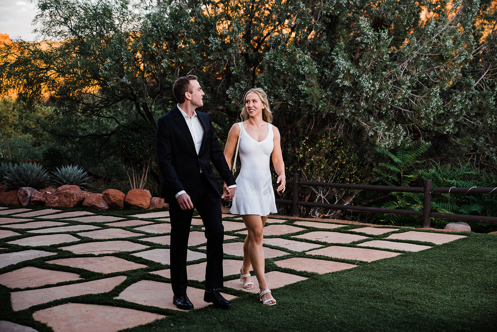 A man in a black suit walks through a manicured garden holding hands with his new fiance after a proposal