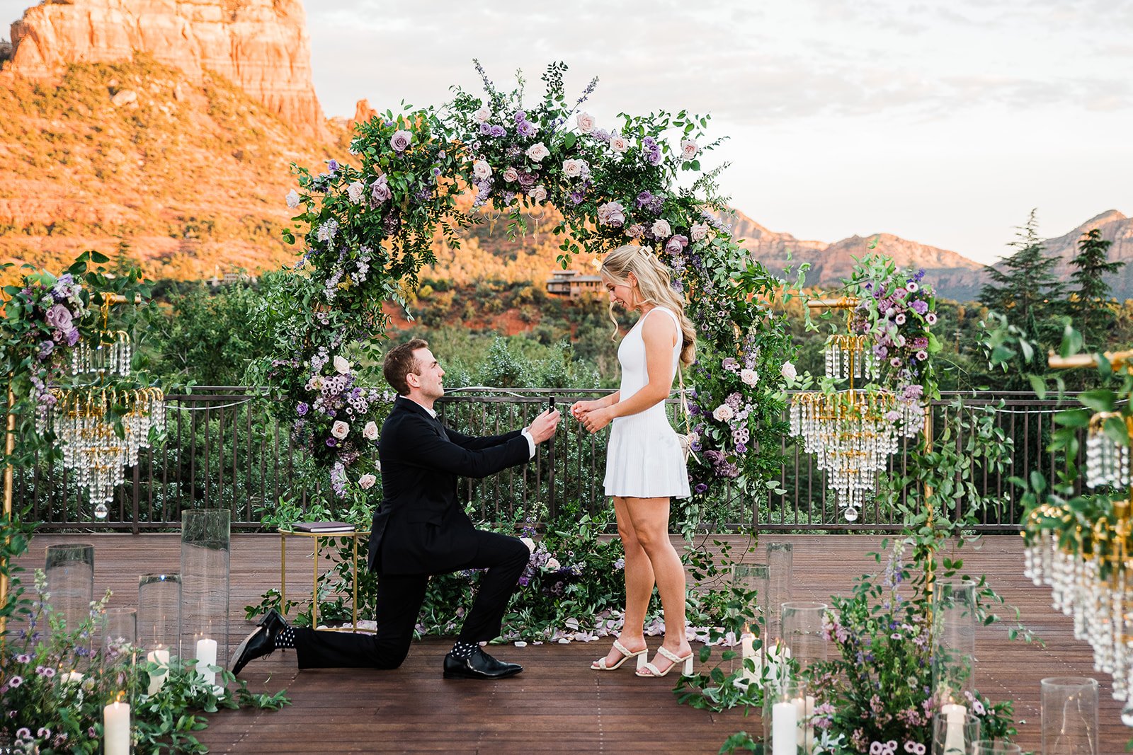 A man in a black suit on one knee proposes with a ring to his girlfriend in a white dress in a mountain park surrounded by romantic florals and decorations