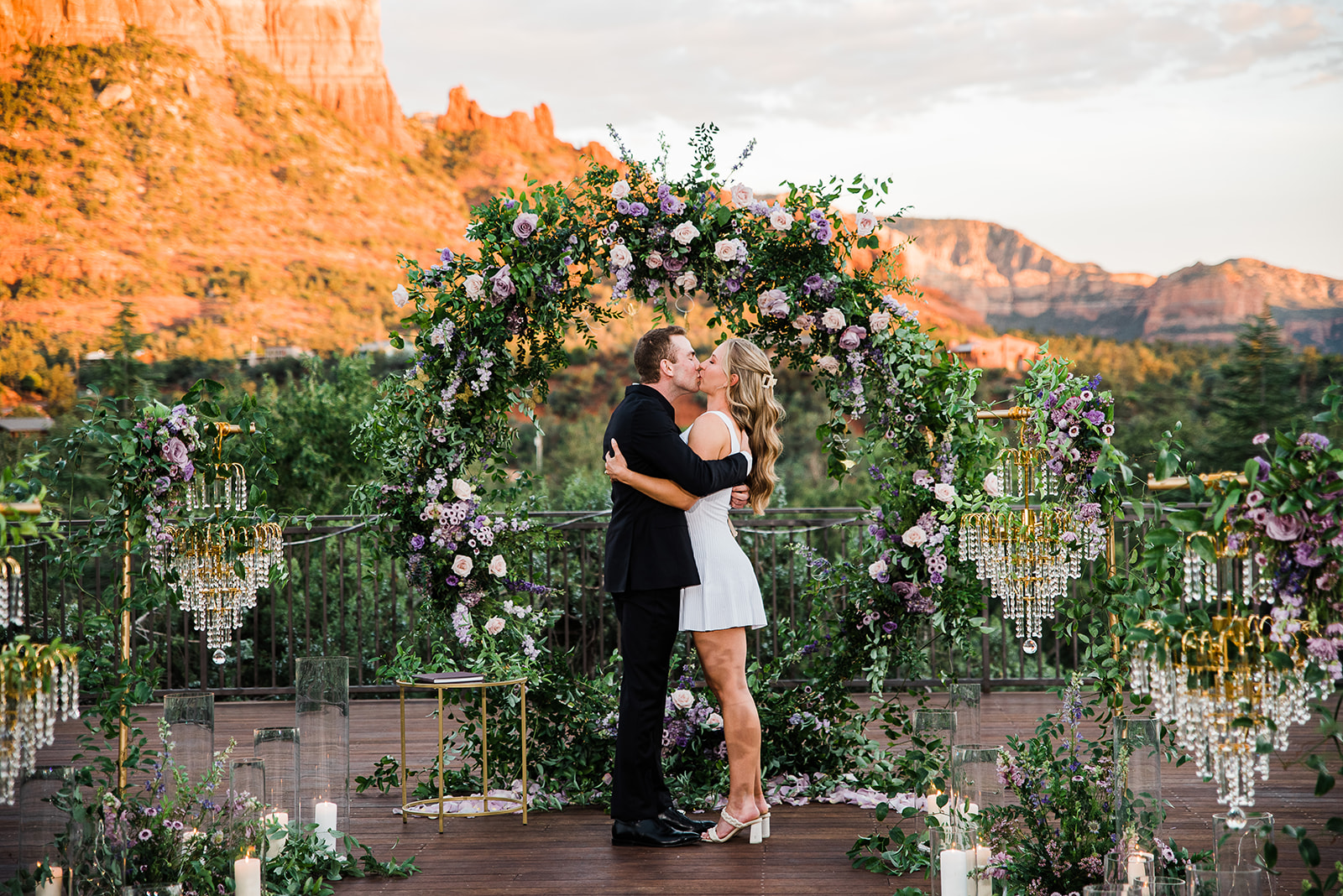 Newly engaged couple kisses in a black suit and white dress at a mountain overlook decorated with chandeliers and purple florals