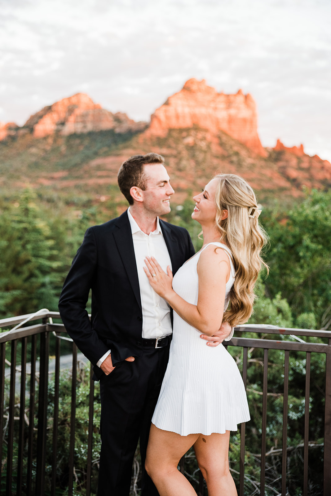 A man in a black suit smilies at his new fiance in a white dress at a desert mountain park after a The Yes Girls proposal