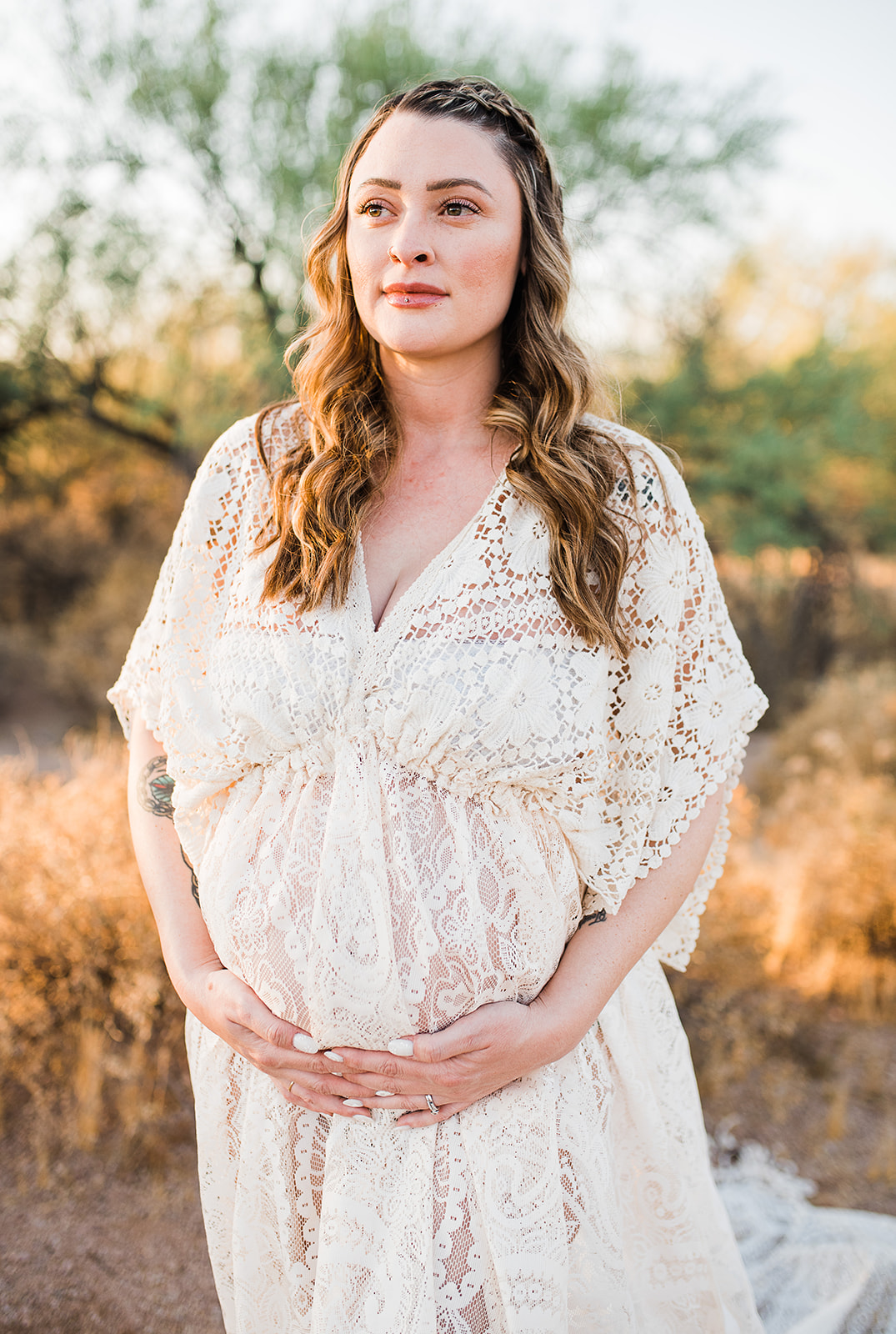 A mom to be smiles into the sunset while holding her bump and standing in a desert wearing a lace maternity gown