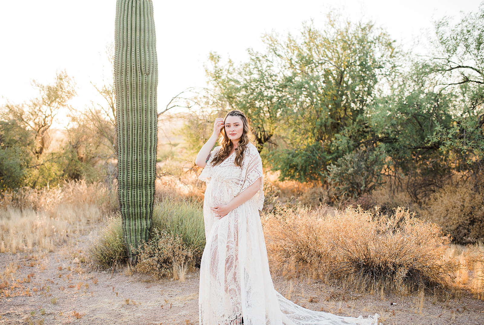 A mother to be pulls back her hair while standing in a desert trail at sunset in a white lace dress after meeting Phoenix midwives
