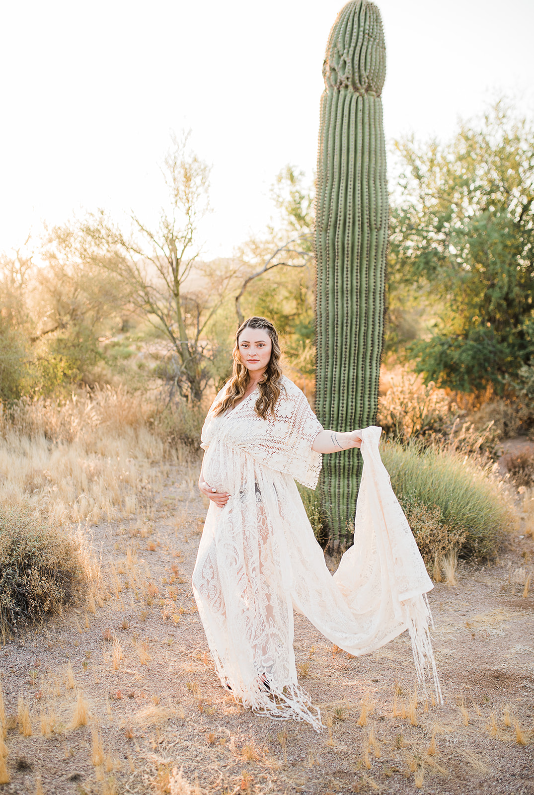A mom to be in a lace maternity dress stands in a desert with a cactus holding her train behind her after meeting Phoenix midwives