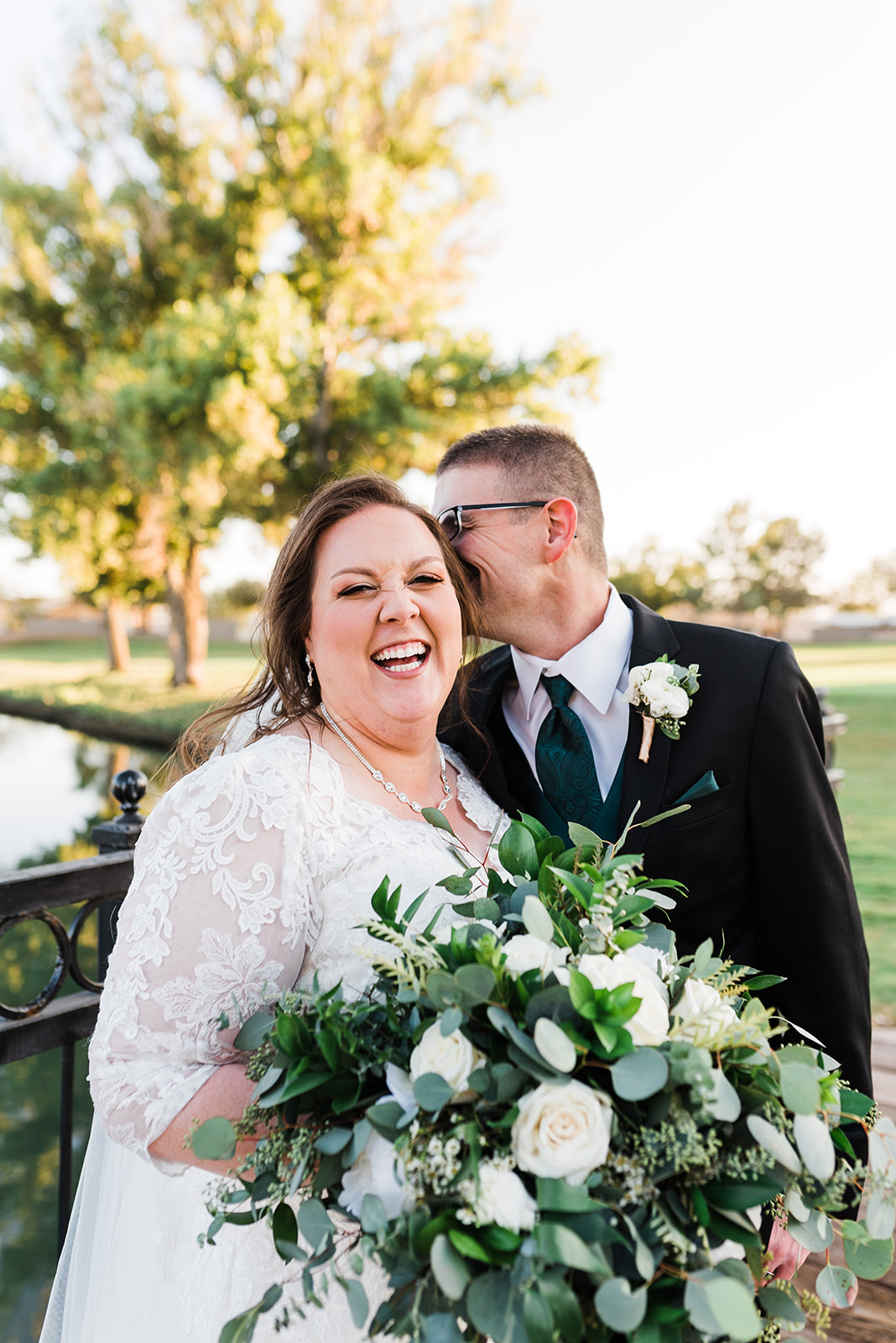 A bride laughs in a lace dress while holding a large white bouquet as her husband whispers in her ear at a Lindsay Grove Wedding