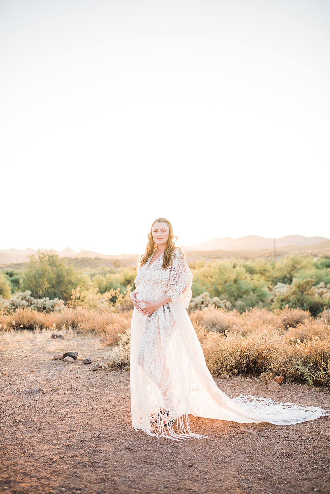 A mother to be holds her bump while standing in a white lace maternity gown in a desert path