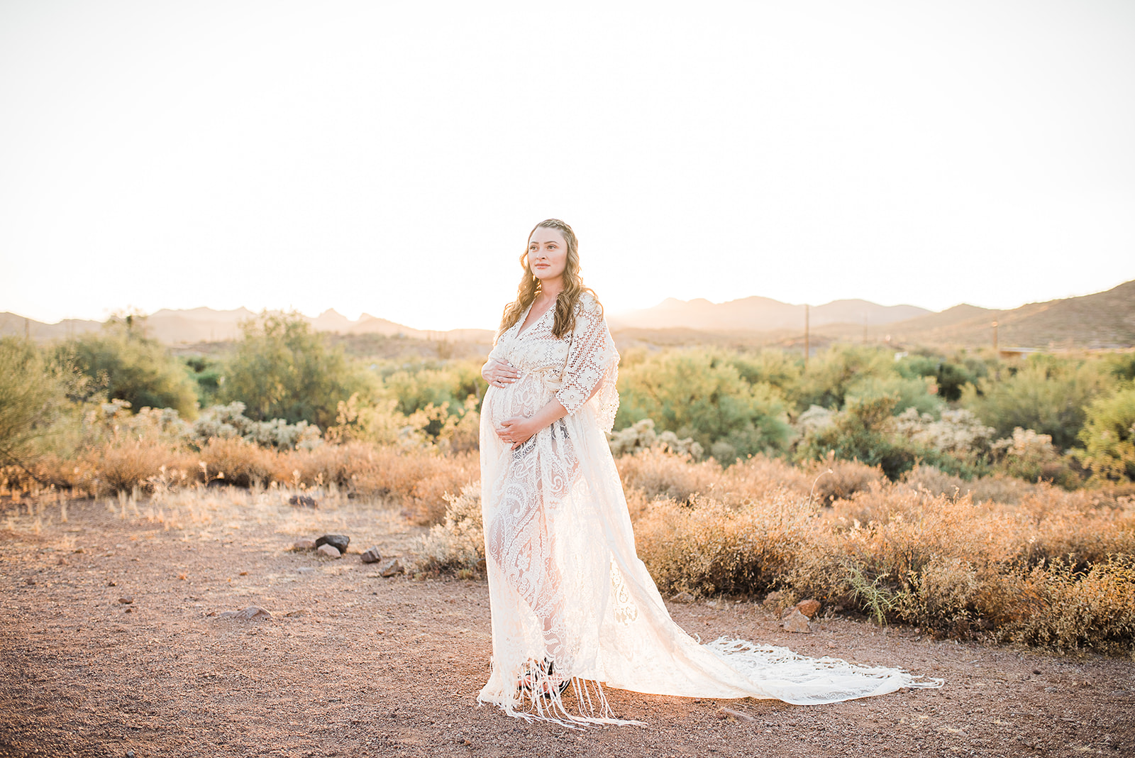 A mom to be in a white lace dress stands in a desert trail at sunset with hands on her bump after visiting Phoenix Doulas