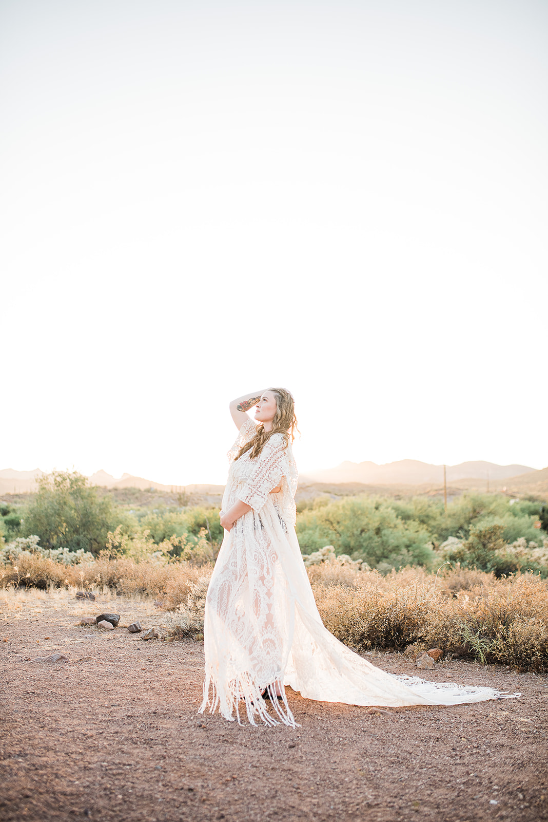 A mom to be in a long white lace dress stands in a desert trail at sunset with a hand in her hair after meeting Phoenix Doulas