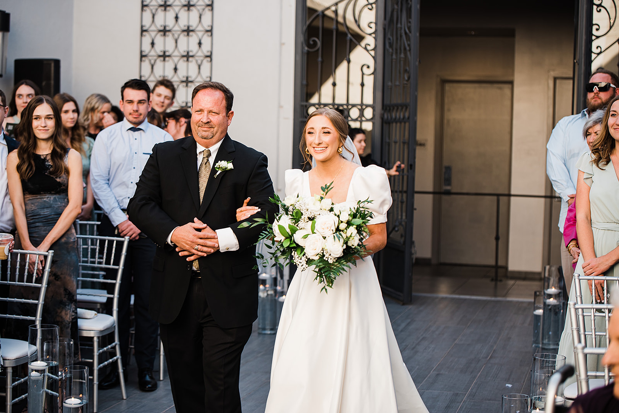A father in a black suit walks his daughter down the aisl in a white silk wedding dress