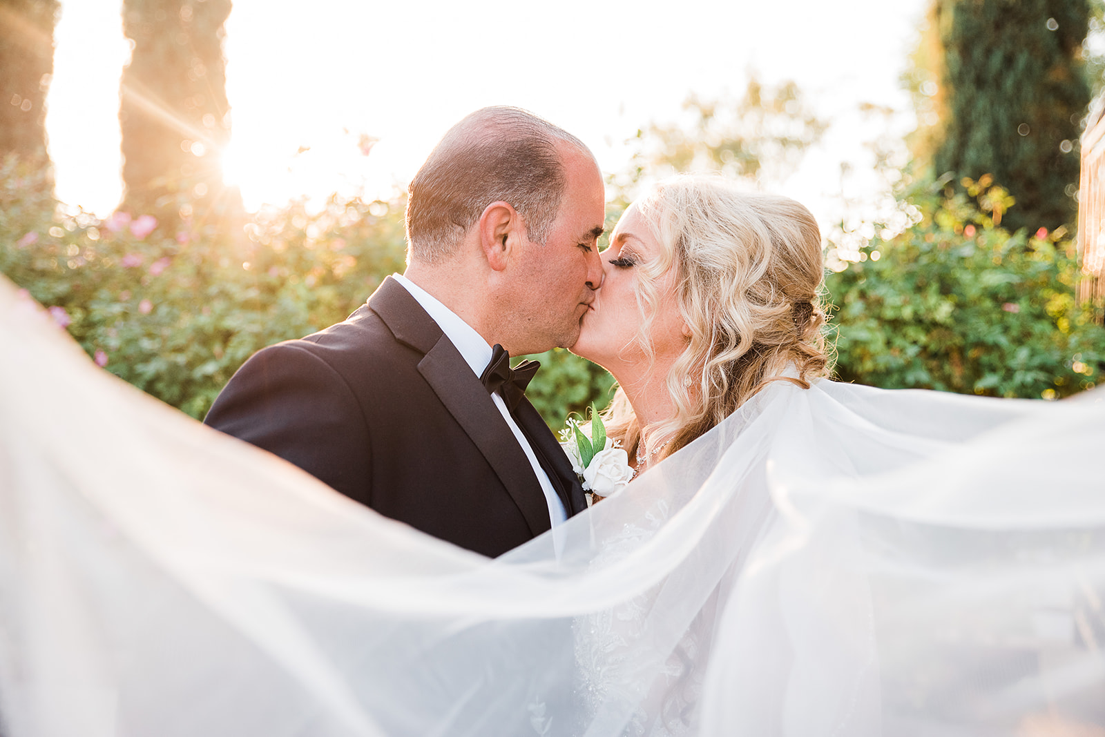 NEwlyweds kiss as the veil flows in the wind at sunset in a garden