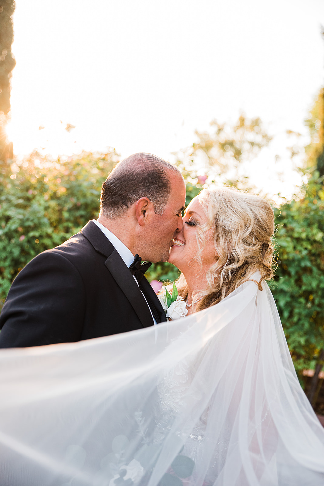 Newlyweds kiss while wrapped in the long veil in a garden at sunset during their Regency Garden Wedding