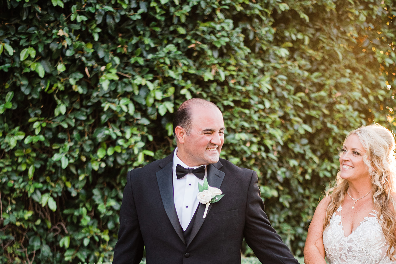 Newlyweds smile big while walking through a garden in a black tux and lace dress at their Regency Garden Wedding