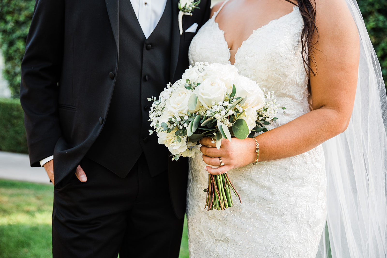 Newlyweds stand in a garden holding the white bouquet in a lace dress and black tuxedo