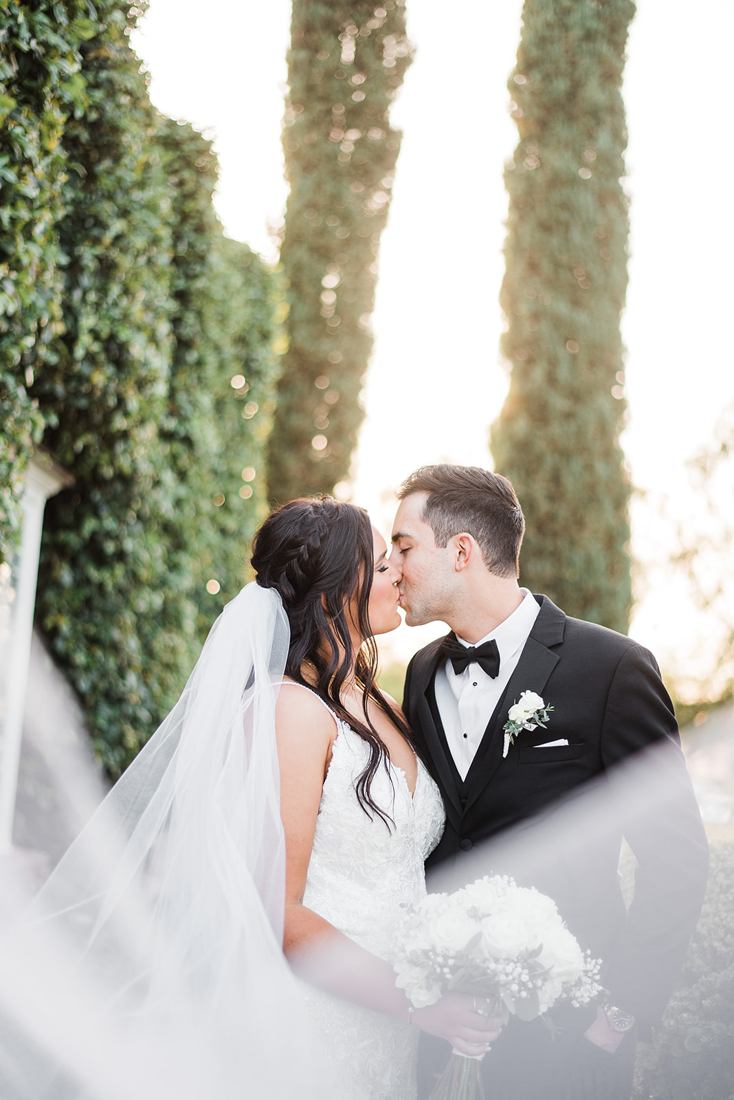 Newlyweds kiss while the veil flows in front of them in a garden at their Venue At The Grove Wedding